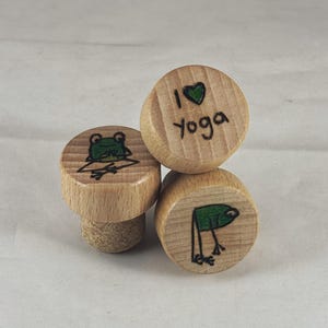 Può includere: Tre tappi per bottiglie in legno con diversi disegni. Un tappo ha una rana verde che medita, un altro ha una rana verde che fa yoga e il terzo ha il testo "Amo lo yoga" con un cuore verde.