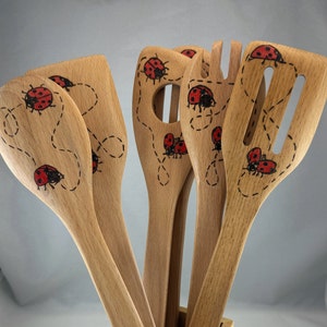 May include: A set of six wooden kitchen utensils, including a spoon, spatula, and slotted spoon. Each utensil is decorated with a red and black ladybug design and a dotted line. The utensils are made of light-colored wood.