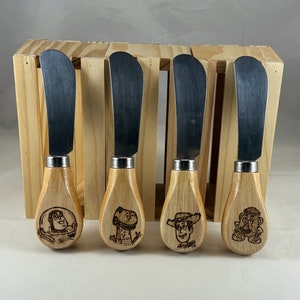 May include: Four wooden butter knives with different cartoon characters engraved on the handles. The characters are Woody, Buzz Lightyear, Rex, and Mr. Potato Head.