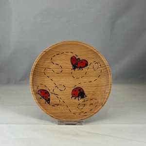 May include: A wooden bowl with a natural wood finish and a design of three red ladybugs with black spots on a white background. The ladybugs are drawn in a simple, cartoon style and are crawling along a path of dotted lines.