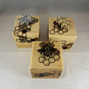 Może przedstawiać: Trzy drewniane pudełka z wzorem plastra miodu i pszczołą na górze. Pszczoły są wykonane z metalu i są srebrne, złote i czarne.