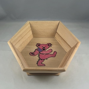 May include: A wooden, hexagonal-shaped bowl with a light brown finish. The interior features a pink dancing bear graphic with a purple scarf. The bowl has a raised base and is likely used for decorative or storage purposes.