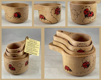 Puede incluir: Conjunto de cuatro tazas medidoras de madera con diseños de mariquitas. Las tazas están anidadas y tienen un acabado de madera marrón. Las mariquitas están pintadas en rojo y negro con puntos blancos. Las tazas están pintadas a mano y son aptas para alimentos.
