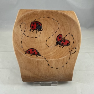 May include: A wooden dish with a natural finish features a design of three ladybugs with black spots and red wings. The ladybugs are drawn with a dark brown line and are surrounded by a dotted line.