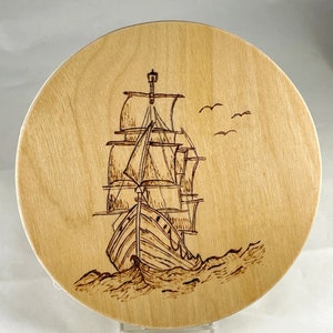 May include: A wooden round plaque with a woodburned image of a sailing ship with billowing sails, navigating through choppy waves. The ship is facing away from the viewer, with the bow of the ship in the foreground.