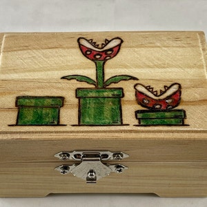 Könnte beinhalten: Eine Holzkiste mit einem Scharnierdeckel und einem Metallverschluss. Die Kiste ist mit einem eingebrannten Bild von drei Piranha-Pflanzen aus der Super Mario Bros.-Videospielserie verziert. Die Pflanzen befinden sich in ihren charakteristischen grünen Töpfen.
