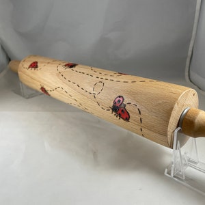 Puede incluir: Un rodillo de madera con un diseño de mariquitas rojas y líneas punteadas. El rodillo está sobre un soporte de acrílico transparente.