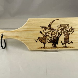 Puede incluir: Una tabla de cortar de madera con un diseño pintado a mano de personajes de Pesadilla Antes de Navidad. Los personajes son Jack Skellington, Sally y Oogie Boogie. La tabla tiene un lazo de colgar negro.