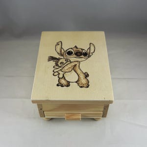Puede incluir: Una pequeña caja de madera de color claro con un cajón. La parte superior de la caja presenta una imagen detallada, grabada en madera, de un personaje de dibujos animados sosteniendo un peluche. El cajón tiene un pequeño tirador rectangular de madera.