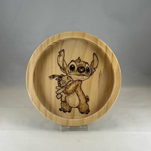 Puede incluir: Un cuenco redondo de madera con un diseño pirograbado del personaje de Disney Stitch abrazando una muñeca. El cuenco está hecho de madera clara y tiene un acabado liso y natural. La obra de arte es detallada y presenta las características distintivas del personaje.