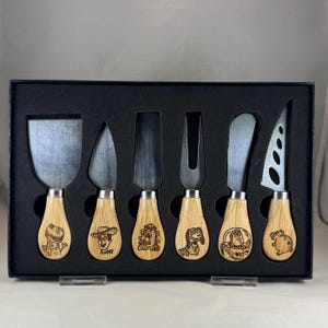 May include: Set of six stainless steel cheese knives with wooden handles. Each handle features a different character from the Toy Story movie franchise. The knives are displayed in a black box.