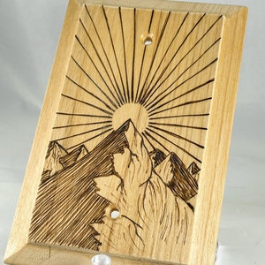 May include: A wooden light switch cover with a woodburned design of mountains and a sun. The design is in shades of brown.