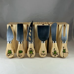 Può includere: Un set di sei coltelli da formaggio con manici in legno e lame in acciaio inossidabile. Ogni manico presenta un'illustrazione diversa di una rana verde, con la scritta "I Yoga" su uno di essi. I coltelli sono disposti in un supporto di legno.