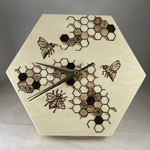 May include: Hexagon-shaped wooden clock with a light wood finish. The clock face features a detailed pyrography design of honeycombs and bees. The clock hands are a dark metallic green. The overall design is rustic and nature-inspired.