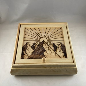May include: A wooden box with a hinged lid and a small knob. The lid features a woodburned design of mountains and a sun with rays.