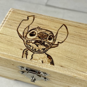 May include: A wooden box with a hinged lid and a metal latch. The top of the box is decorated with a wood-burned image of Stitch from the Disney movie Lilo & Stitch.