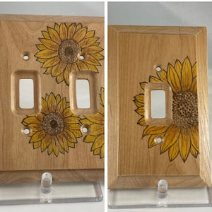 Puede incluir: Dos tapas de interruptor de luz de madera con un diseño decorativo de girasol. Las tapas son de color marrón claro con un acabado de madera natural. Los girasoles son amarillos con centros marrones.
