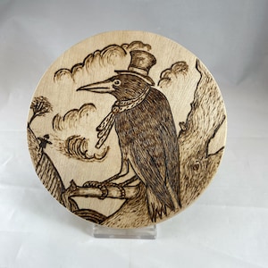 May include: A woodburned round plaque with a detailed image of a crow wearing a top hat and scarf, perched on a branch with a cloudy sky background.