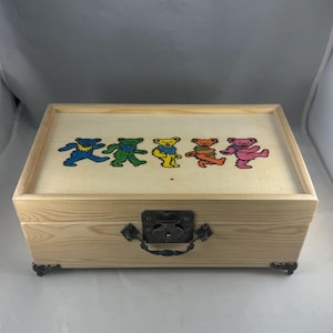 以下が含まれることがあります： ヒンジ付きの蓋と金属製の留め金が付いた木製の箱。蓋には、青、緑、黄、オレンジ、ピンクの5匹の踊るクマのカラフルなイラストが描かれています。箱には金属製のコーナーアクセントが付いています。