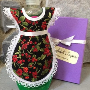 May include: A black and green dish soap bottle with a white lace and red berry print fabric cover. The cover has a white ribbon and a white lace trim. The bottle is sitting on a stone surface next to a purple box with a white ribbon and a white tag that says "Thoughtful Elegance".
