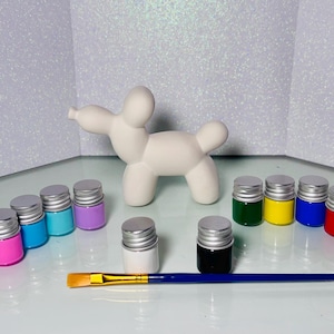 Puede incluir: Una figura de perro globo de cerámica blanca con un juego de pinturas coloridas y un pincel. Los colores de pintura incluyen rosa, azul, turquesa, morado, blanco, negro, verde, amarillo, azul y rojo. Los botes de pintura tienen tapas plateadas.
