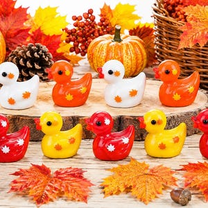 Pode incluir: Uma coleção de nove patinhos de borracha pequenos nas cores branca, laranja, amarela e vermelha, cada um decorado com padrões de folhas de bordo contrastantes. Os patos estão dispostos em duas filas sobre uma superfície de madeira e folhas.