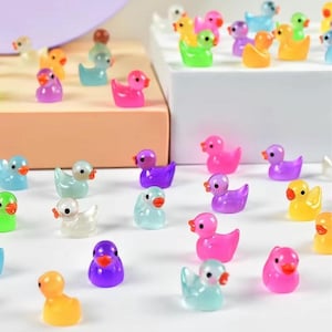 Puede incluir: Una colección de pequeñas figuras de patos coloridos en varios tonos de rosa, morado, amarillo, verde y azul. Cada pato tiene un pico naranja y está dispuesto sobre una superficie blanca, creando una escena lúdica y caprichosa.