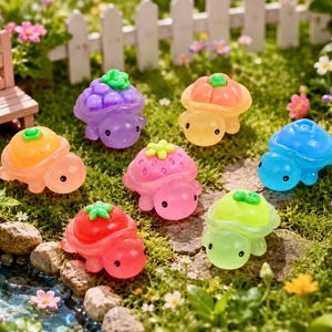 Puede incluir: Una colección de siete figuras de tortugas translúcidas, cada una con un caparazón en forma de fruta. Las tortugas son de color morado, naranja, azul, rosa, rojo y verde claro, con ojos negros y tallos verdes en la parte superior.