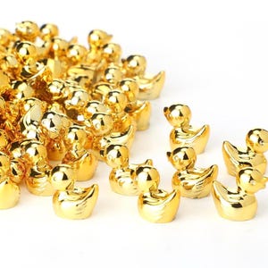 Puede incluir: Un conjunto de pequeñas figuras de patos dorados y brillantes. Los patos están agrupados, con algunos dispersos en primer plano. Las figuras tienen una superficie lisa y reflectante, sobre un fondo blanco. Los patos son un objeto decorativo.