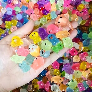 Puede incluir: Una mano sosteniendo una colección de amuletos coloridos y translúcidos con forma de animales. Los amuletos incluyen patos, ranas, delfines y otras criaturas en tonos de amarillo, rosa, azul, verde y morado. El fondo está lleno de amuletos similares.