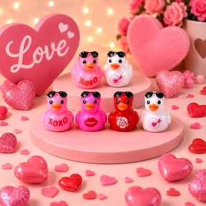 Könnte beinhalten: Eine Sammlung von sechs kleinen Quietscheentchen auf einer runden rosa Plattform. Jede Ente trägt eine schwarze Sonnenbrille und zeigt einzigartige Motive wie den Schriftzug XOXO, eine rote Rose, ein Herz mit Pfeil sowie ein Paar rote Lippen.