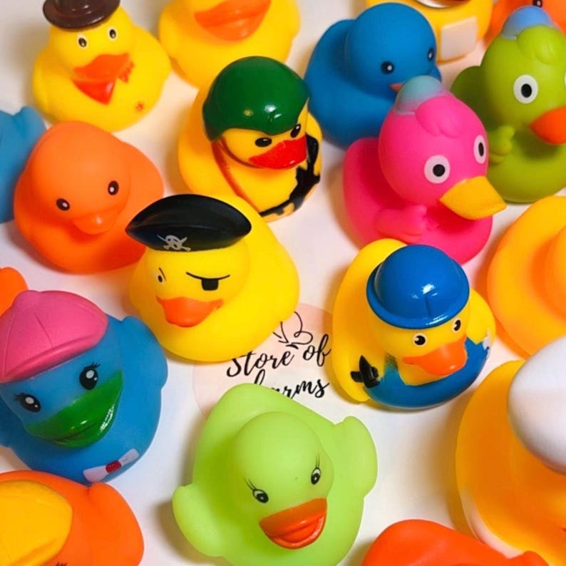 May include: A collection of colorful rubber ducks in various designs, including a pirate duck with an eye patch, a duck wearing a brown hat, a pink duck, a blue duck with a pink cap, and a green duck, arranged together on a white surface.