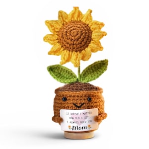 May include: A crocheted sunflower with yellow petals and a brown center, green leaves, and a brown pot. The pot has a smiling face and holds a sign that reads "It doesn't matter how old I get, I always need you Mom."