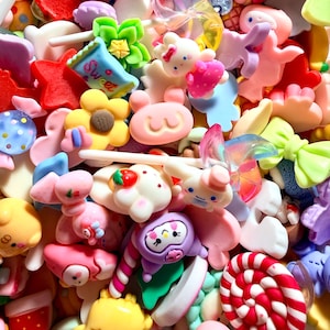 Puede incluir: Un colorido surtido de pequeños dijes decorativos de resina en varias formas, incluyendo flores, estrellas, animales y dulces. Los dijes son en tonos de rosa, amarillo, rojo, azul y verde, algunos con purpurina y la palabra "Sweet".