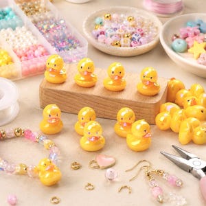 May include: A collection of small yellow rubber duck charms, assorted colorful beads in a plastic organizer, a heart charm, and jewelry findings including earring hooks and jump rings, with a partially assembled beaded bracelet.