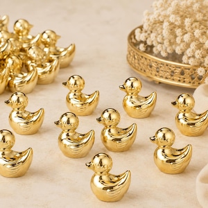 Puede incluir: Una colección de pequeñas figuras de patos de color dorado brillante. Los patos tienen la forma de patitos de goma clásicos con superficies lisas y reflectantes. Varios patos individuales están dispuestos en primer plano, junto a un grupo.