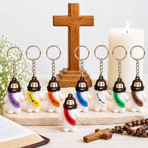 May include: A collection of eight keychains featuring a cartoon figure of Jesus with brown hair and a beard, wearing a white robe with a colored sash printed with the text Jesus + you. The sashes are purple, yellow, orange, red, blue, white, green, and brown.