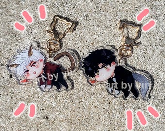 May include: Two acrylic keychains featuring anime-style characters with cat ears and tails. One character has white hair and a red jacket, the other has black hair and a black suit. Both keychains have gold-colored cat-shaped clasps. The text "art by yii" is visible.