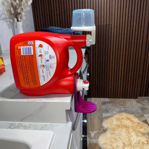May include: A red Tide laundry detergent bottle with a purple dispenser attachment. A clear blue measuring cup is attached to the dispenser. The bottle is on a white countertop.