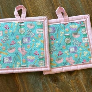 May include: Two pink and white quilted pot holders with a blue background and a pattern of kitchen tools, cupcakes, and the word "Recipe".