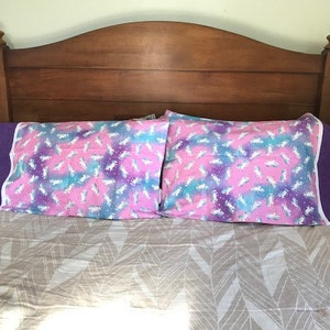 May include: Two pink and purple pillowcases with a white unicorn pattern on a bed with a brown headboard and a beige patterned duvet.