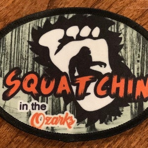 May include: Oval patch with a black border featuring a white Bigfoot footprint and silhouette. The text "Squatchin in the Ozarks" is in orange and black. The background has a green and white forest pattern.