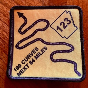 May include: A yellow square patch with black stitching around the edges. The patch has a black and purple winding road design with the text "199 curves next 64 miles" and a white diamond with the number "123" inside.