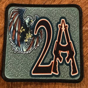 May include: A gray square patch with a black border. The patch features a stylized American bald eagle with red, white, and blue feathers. The eagle is holding a rifle in its talons. The number "2" and the letter "A" are in black with an orange outline. The letter "A" is formed by two rifles.