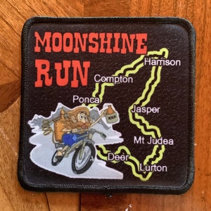 May include: A black patch with a white cartoon illustration of a person riding a motorcycle with a jug of moonshine. The text "Moonshine Run" is in red and white. The patch also includes a map of a route with the names of towns: Compton, Ponca, Harrison, Jasper, Mt Judea, Deer, and Lurton.