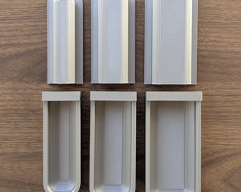 May include: Six light gray, rectangular, open-top containers of varying sizes are arranged on a wooden surface. Each container has a rounded top and a vertical, textured panel in the center. The containers appear to be made of plastic.