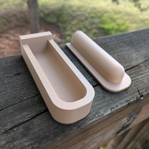 May include: Two beige, 3D-printed objects sit on a weathered wooden surface. One is a rectangular trough with rounded corners, and the other is a rounded, cylindrical shape on a flat base. The objects appear to be a mold and its corresponding part.