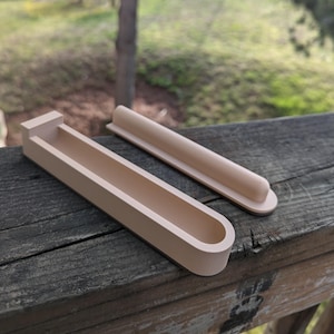May include: Two beige, 3D printed, rectangular-shaped holders with a rounded edge. The larger holder has a curved, open top and the smaller holder is solid.
