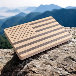 Puede incluir: Un objeto rectangular de color beige con una superficie texturizada, que presenta un diseño detallado de la bandera estadounidense en gris oscuro. La bandera incluye estrellas y rayas, sobre un fondo de montañas.