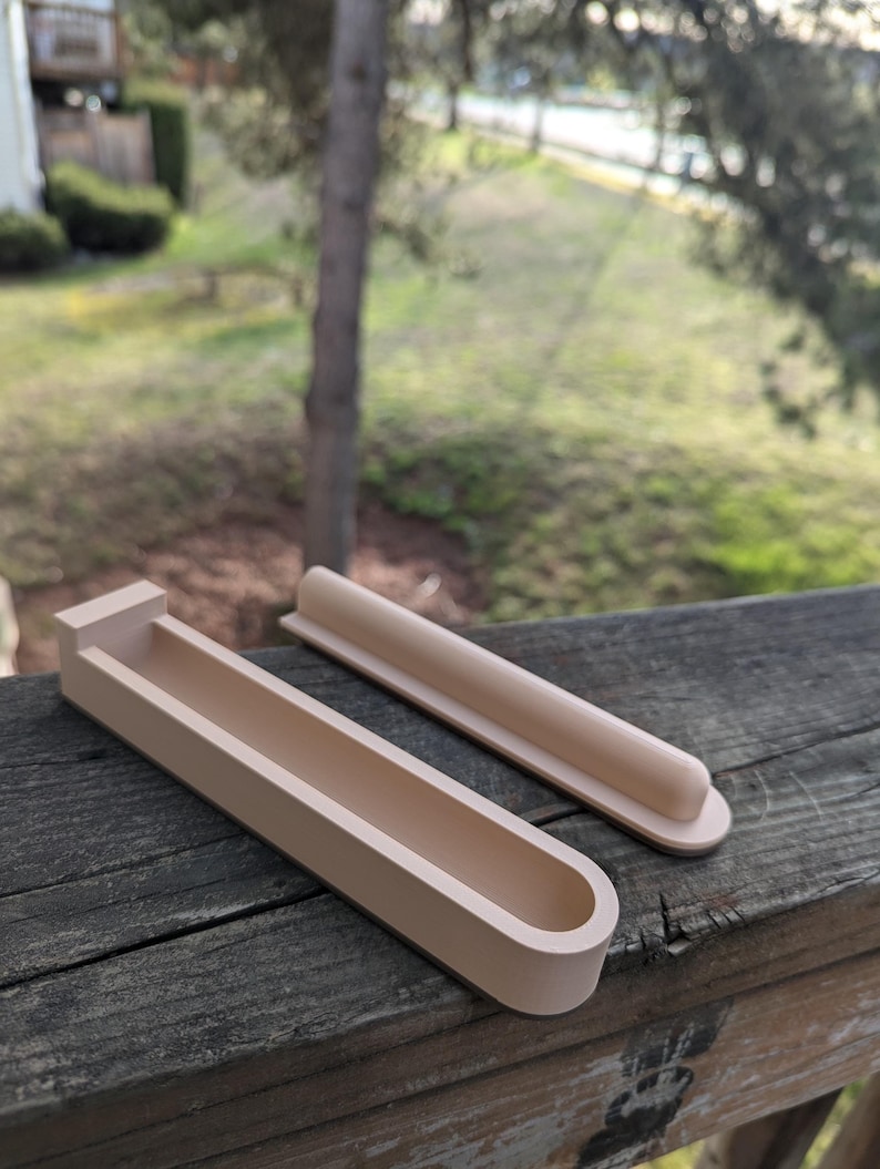 May include: Two beige, 3D printed, rectangular-shaped holders with a rounded edge. The larger holder has a curved, open top and the smaller holder is solid.